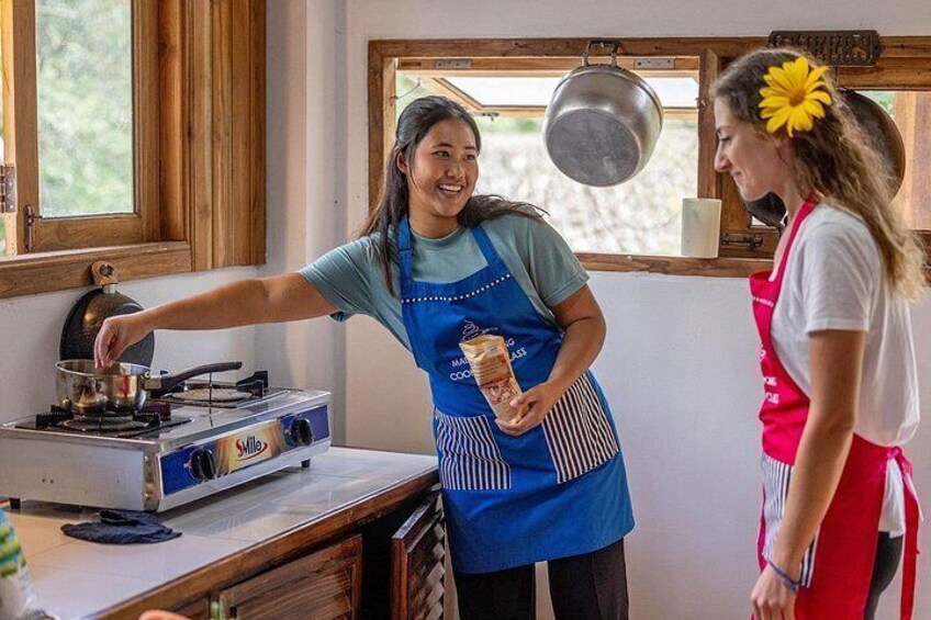 A person cooking on a stove, another person standing nearby, and a window with a view of greenery. 2