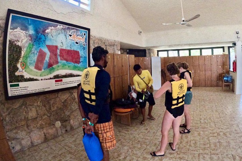 A group of people in a room with a large map on the wall, wooden lockers, and a ceiling fan. 2