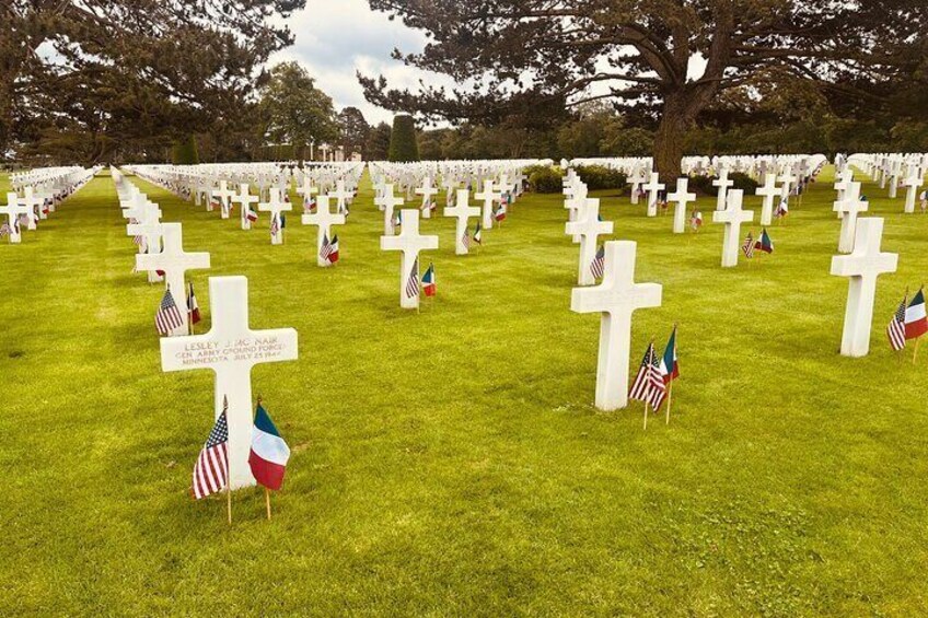 Un cimetière où l’on peut voir de nombreuses croix blanches et des drapeaux, dont les drapeaux américain et français. 3