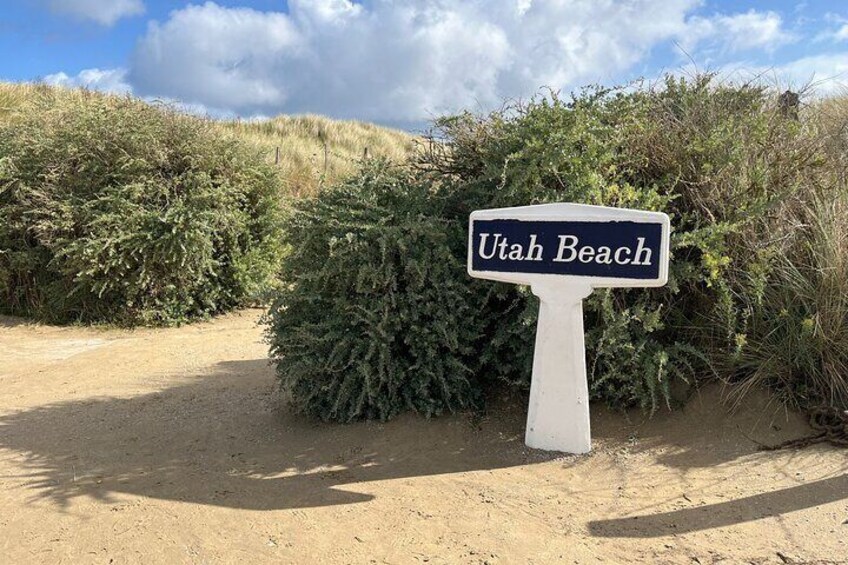 Un panneau indiquant « Utah Beach », placé dans un environnement naturel avec du sable et de la végétation. 2