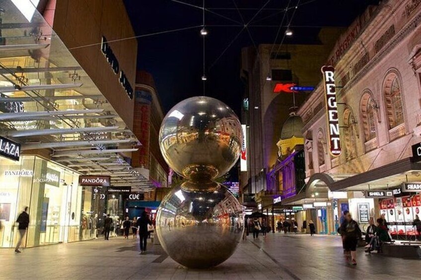 A shopping mall with a large spherical sculpture, storefronts, and a historic building in the background. 1