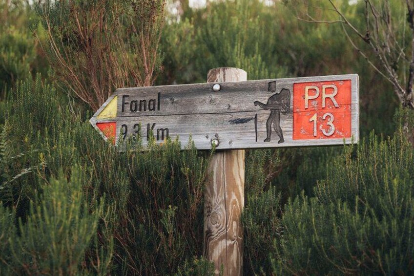 A wooden trail sign with a figure hiking, pointing towards 'Fanal' 93 km away, and a red section labeled 'PR 13'. 2