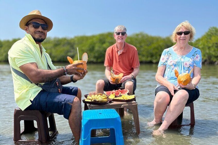 Three people sitting on stools in shallow water, holding coconuts and a plate of food. 4