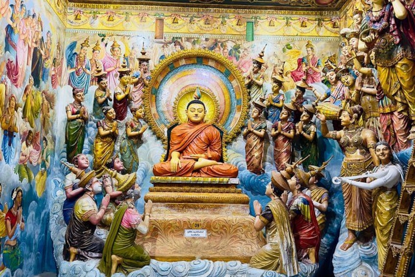 A large Buddha statue surrounded by numerous figures in traditional attire, set against a backdrop of detailed murals. 1
