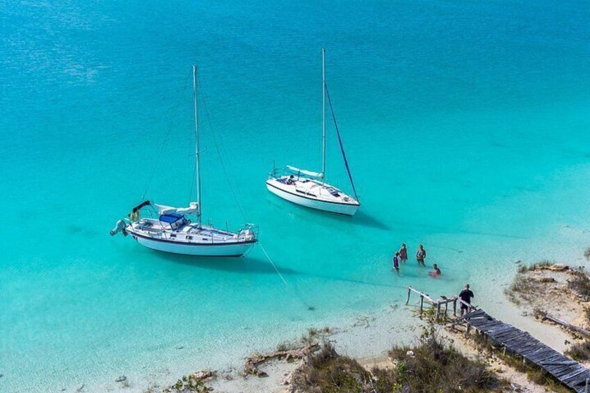 Two sailboats anchored near a beach with people walking on the shore. 1