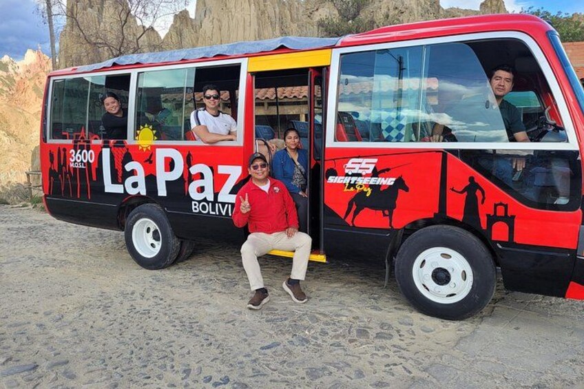 En röd och svart buss med texten ”La Paz” och ”3600 meter” på sidan, parkerad på en sandig yta. 1