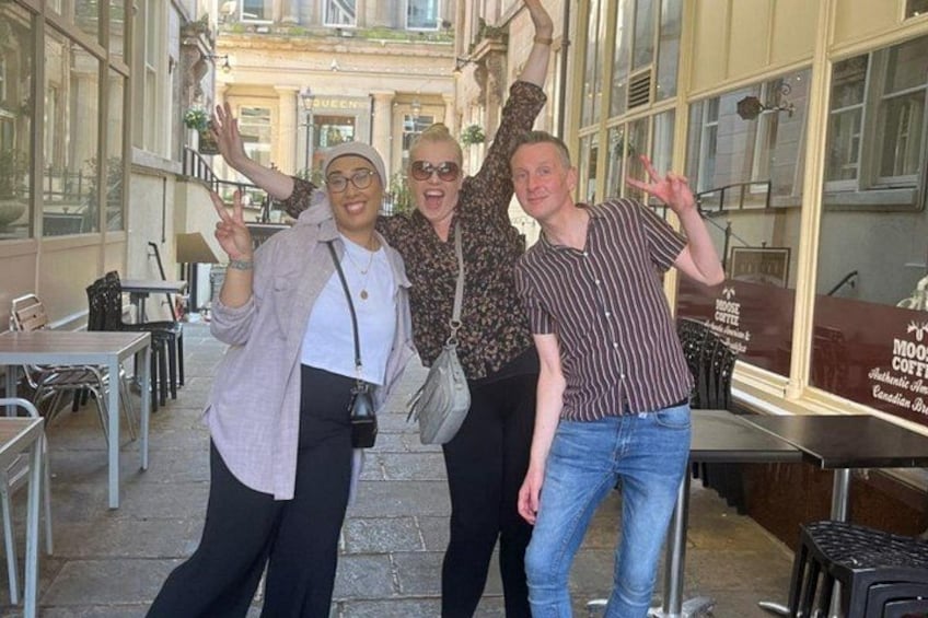 Three people posing in an outdoor cafe setting with tables and chairs, and a sign for a coffee shop in the background. 4