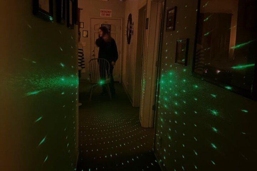 En svagt upplyst korridor med gröna, laserliknande ljus, en person som står vid en dörr och en stol. 3