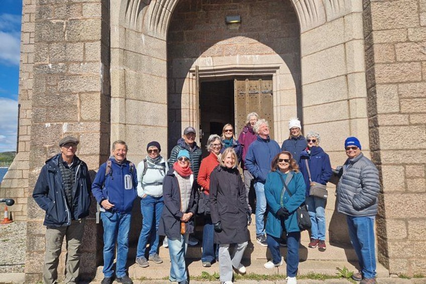 Un groupe de personnes se tient devant un bâtiment en pierre, dont l’entrée est surmontée d’un arc. 4
