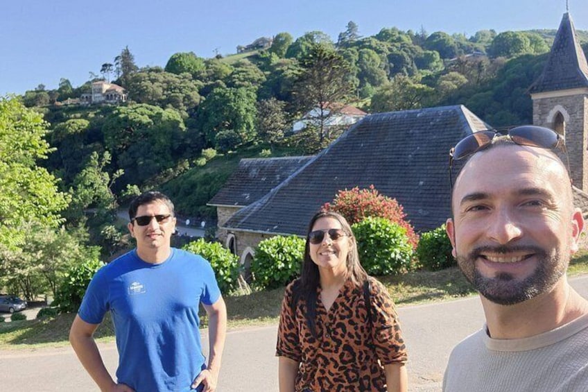 Three people standing on a road with a church and greenery in the background. 3