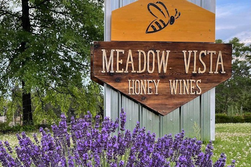 A wooden sign with 'MEADOW VISTA HONEY WINES' written on it, surrounded by purple flowers. 2