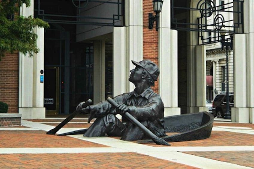 A bronze statue of a man rowing a boat, located on a paved area with brick and stone tiles, in front of a building with columns and a metal railing. 3
