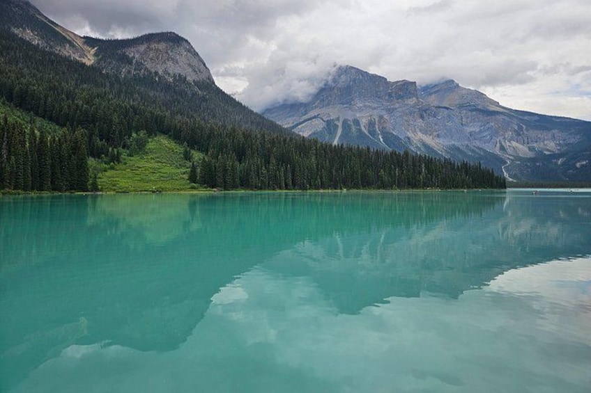 A calm lake with clear turquoise water, surrounded by dense green forests and mountains under a cloudy sky. 2