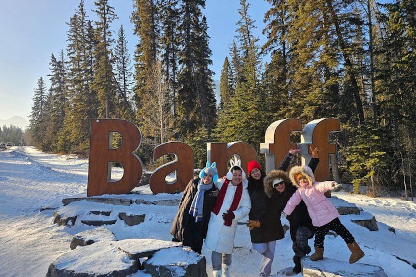 A group of people posing in front of large wooden letters spelling 'Banff' with snow-covered ground and trees in the background. 4