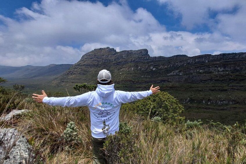 Una persona de pie con los brazos extendidos en una colina cubierta de hierba, con vistas a un paisaje montañoso. 1
