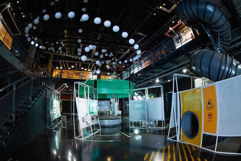 An indoor exhibition space with a high ceiling, industrial lighting, and a central green structure. 5