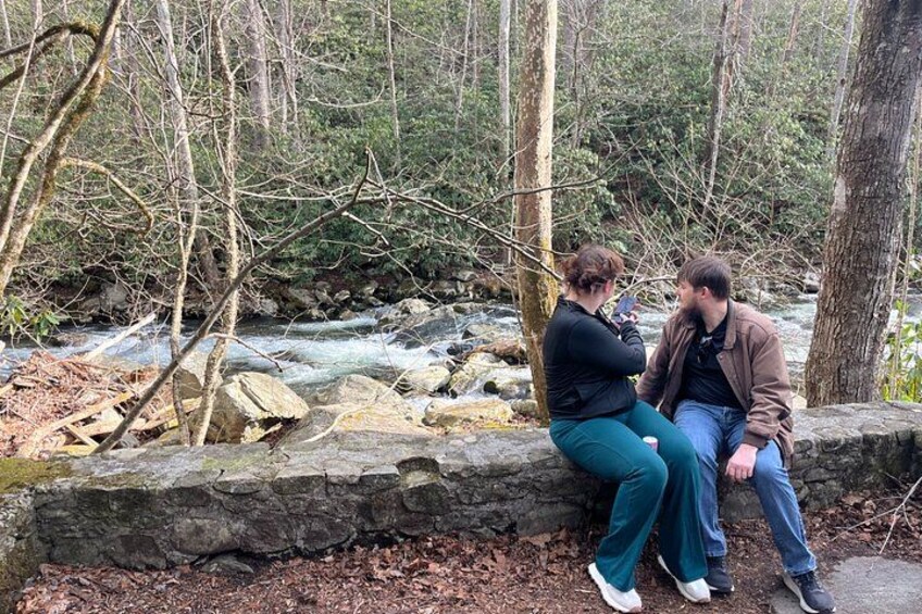 Dos personas sentadas en un borde de piedra junto a un río que fluye en un área boscosa. 3