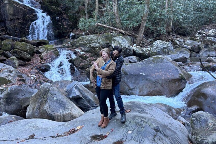 Dos personas de pie sobre rocas junto a una cascada en un área boscosa. 1