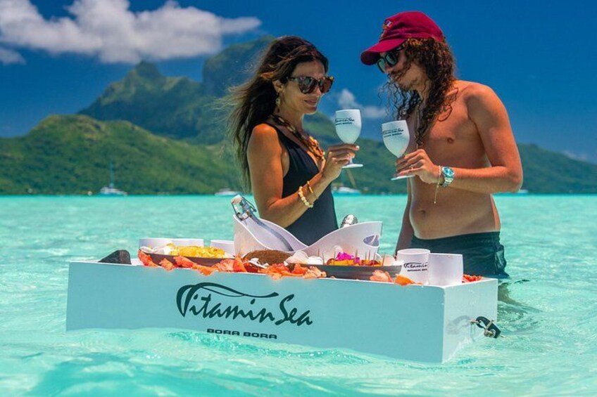 Dos personas en un bote con una caja etiquetada 'VitaminSea' en un entorno tropical. 2