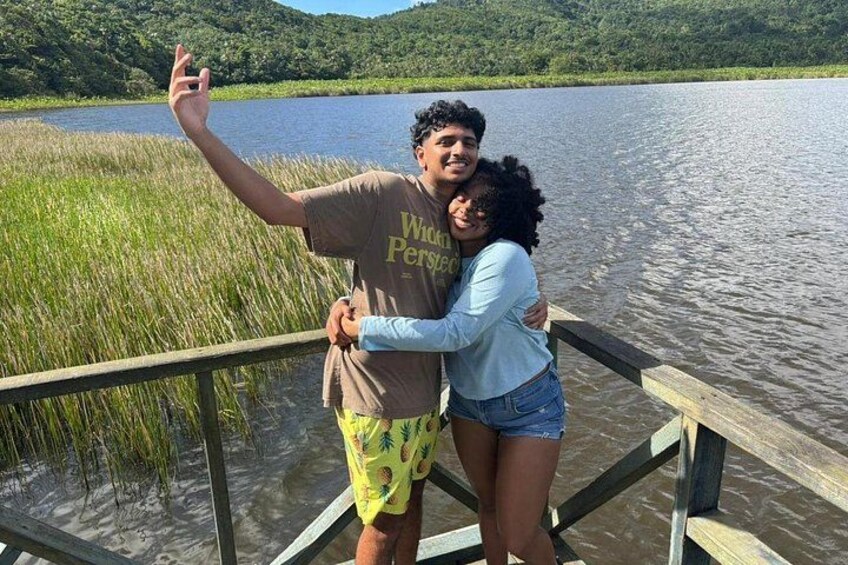 A person taking a selfie on a wooden dock by a lake with tall grass and hills in the background. 5