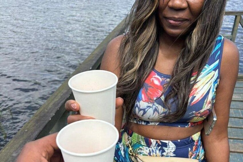 A person holding two paper cups, wearing a colorful floral top, standing on a wooden dock by water. 2