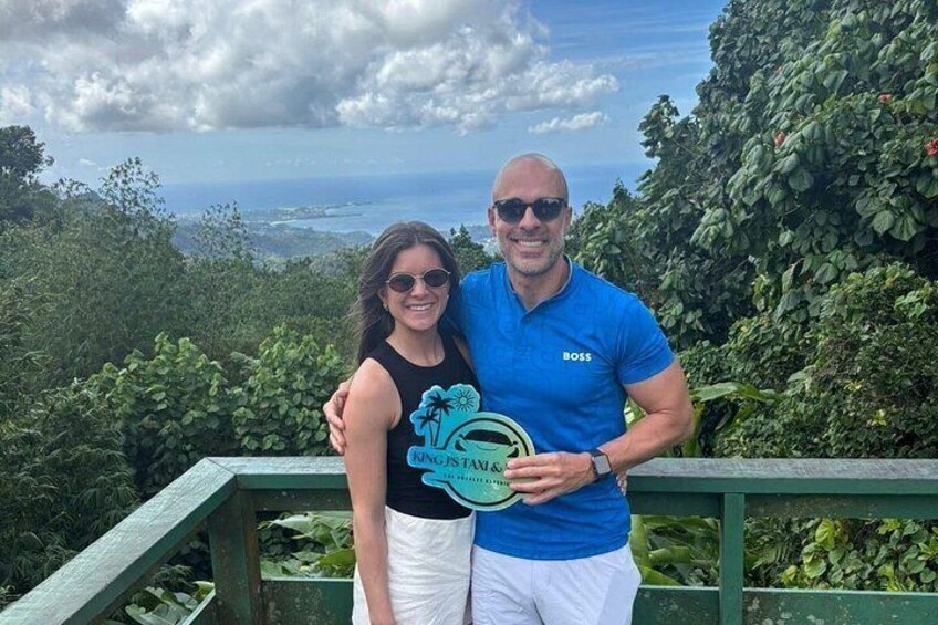 A man and a woman standing on a green railing with a scenic view of the ocean and vegetation in the background. 4