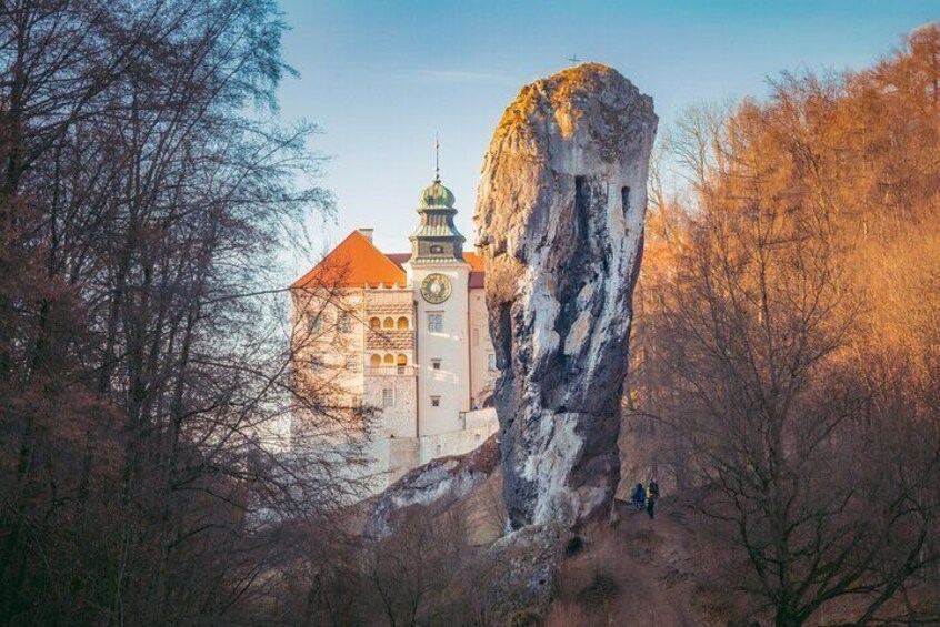 A castle with a clock tower, surrounded by trees and a large rock formation. 1