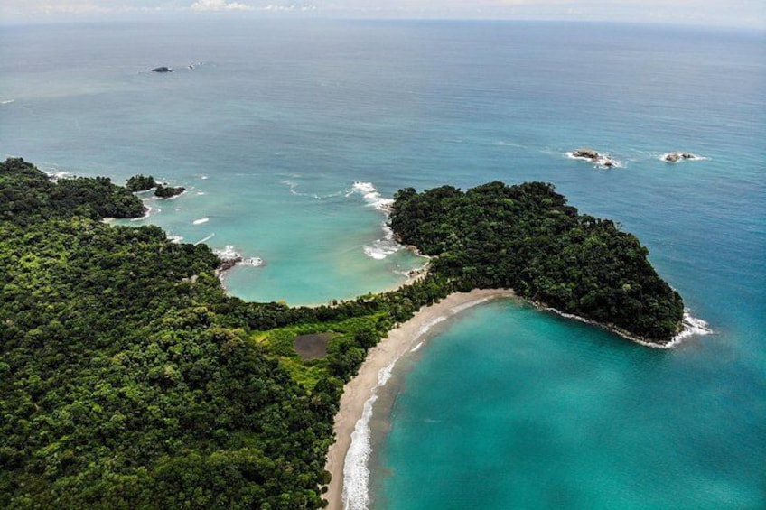 An aerial view of a tropical island with a curved shoreline, dense green forest, and clear turquoise waters. 1