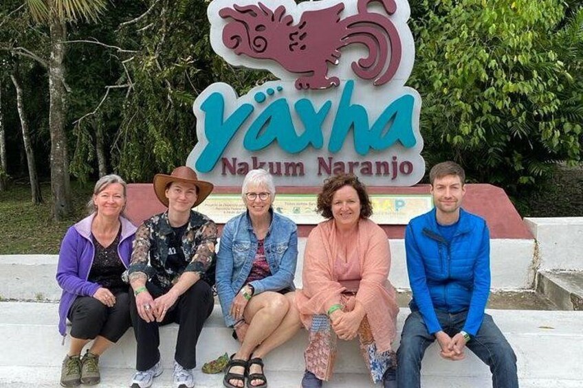 A group of people sitting on steps in front of a sign that reads 'Yayha Naturum Naranjo.' 1