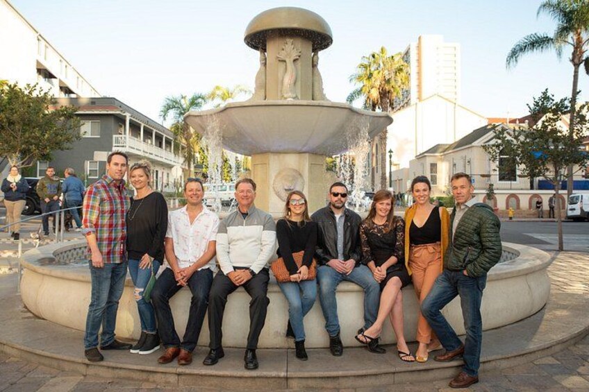 A group of people sitting around a fountain in an urban setting with buildings and palm trees in the background. 2