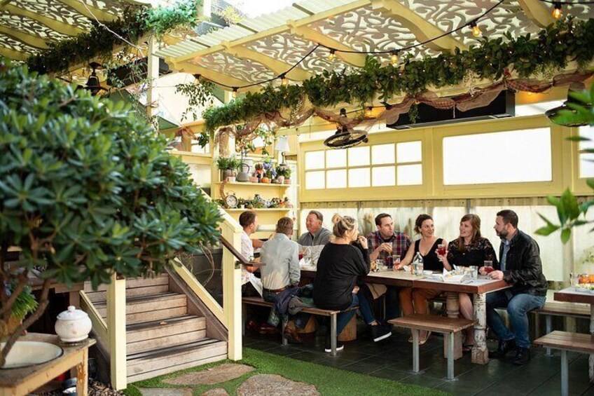An outdoor patio with wooden tables and benches, surrounded by greenery and a decorative trellis. 4