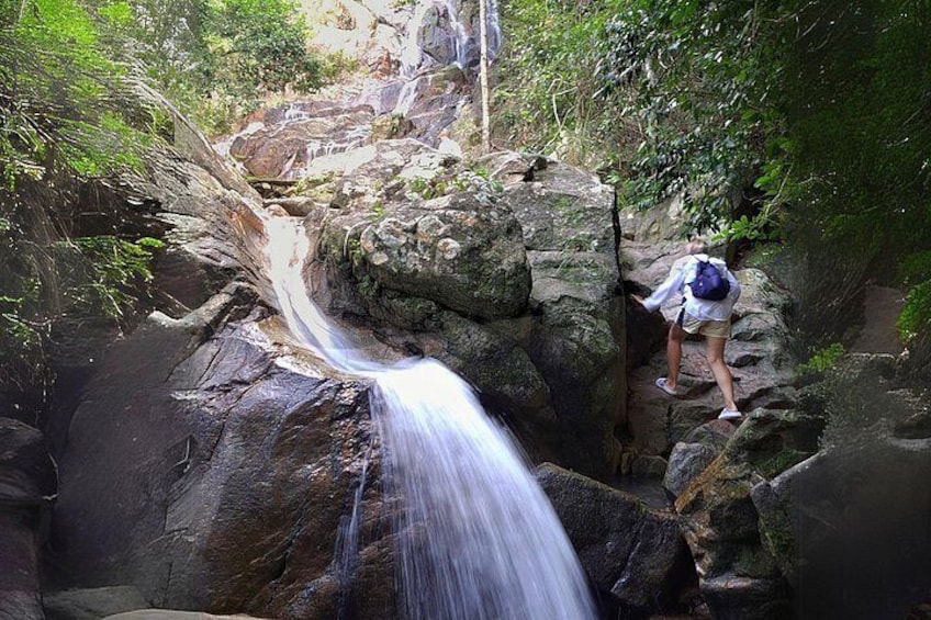 Una cascada que se precipita sobre rocas, una persona escalando un sendero pedregoso y vegetación exuberante al fondo. 2