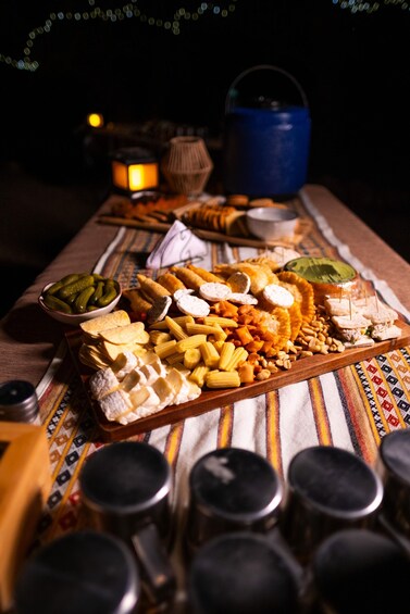 A wooden tray filled with various cheeses, crackers, and pickles, accompanied by a blue kettle and a lantern on a table with a striped cloth. 4