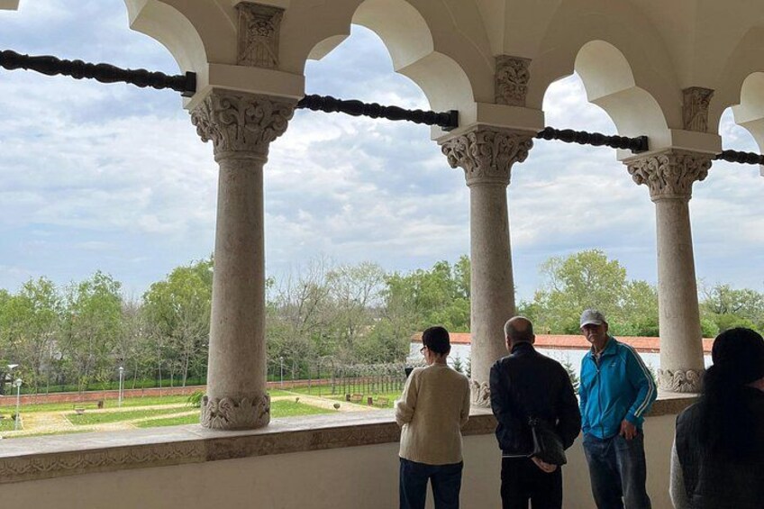 A covered outdoor area with columns, a view of a park with trees and a fence, and people standing and looking out. 4