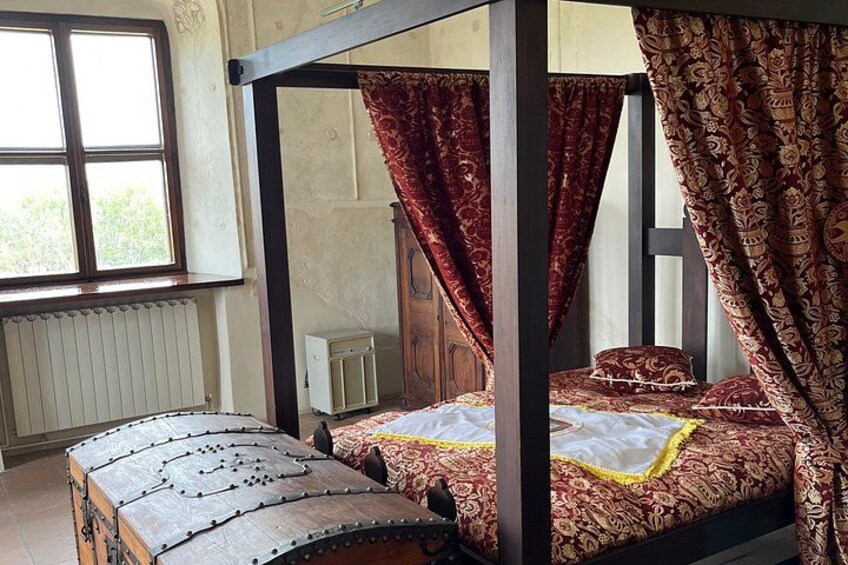 A four-poster bed with red and gold bedding, a wooden chest, and a window with a view of greenery. 2