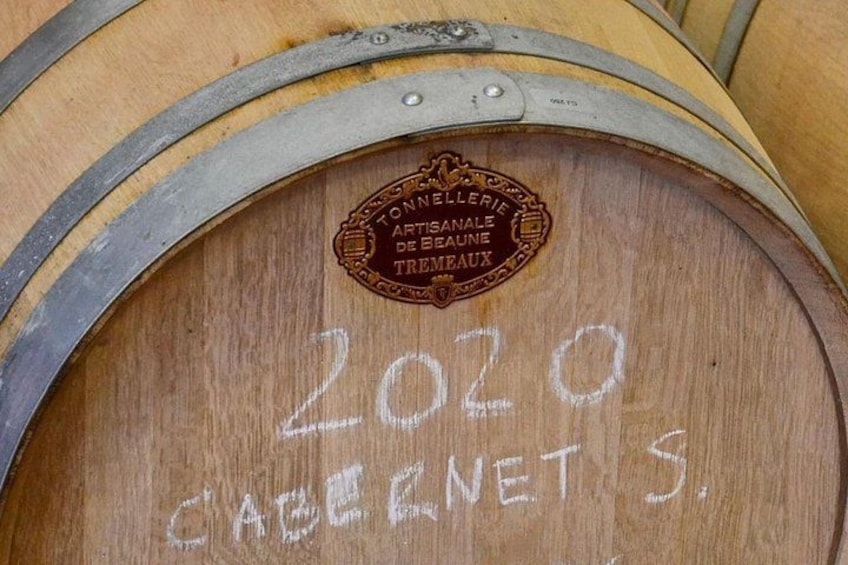 A wooden barrel with a metal band, a label with '2020' and 'CABERNETS' written on it, and a logo with 'TONNELERIE ARTISANALE DE BEAUNE TRAMEAUX'. 2