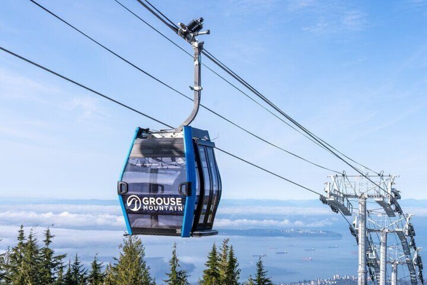 En linbana med texten GROUSE MOUNTAIN som färdas längs en vajer, med ett snötäckt landskap och träd nedanför. 1