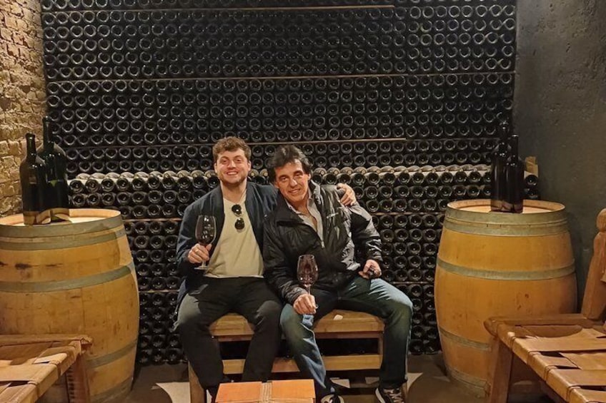 Due persone sedute su una panca in una cantina vinicola, con botti e una parete di bottiglie di vino. 4