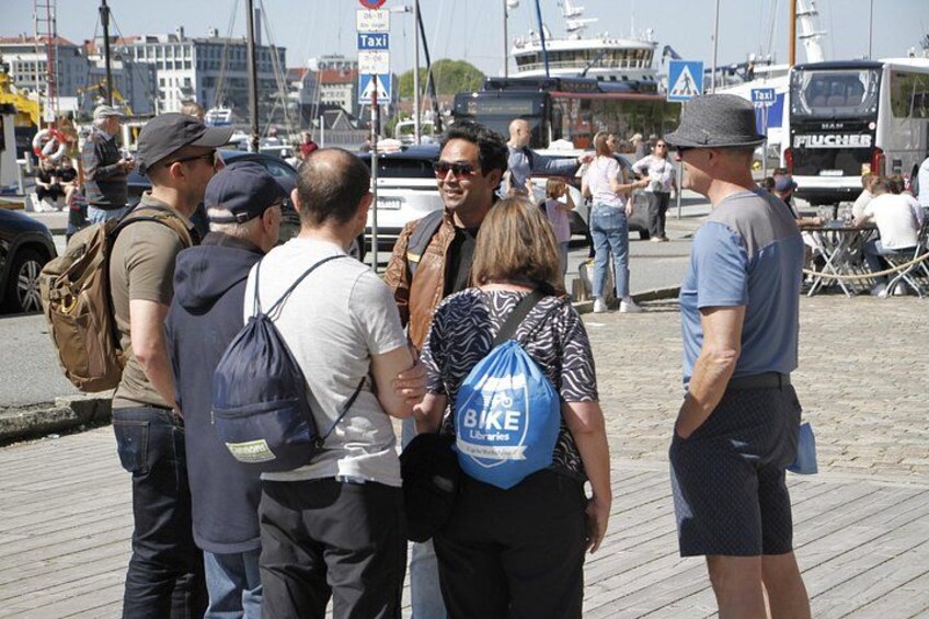 A group of people standing on a wooden boardwalk near a waterfront with boats and a bus in the background. 2