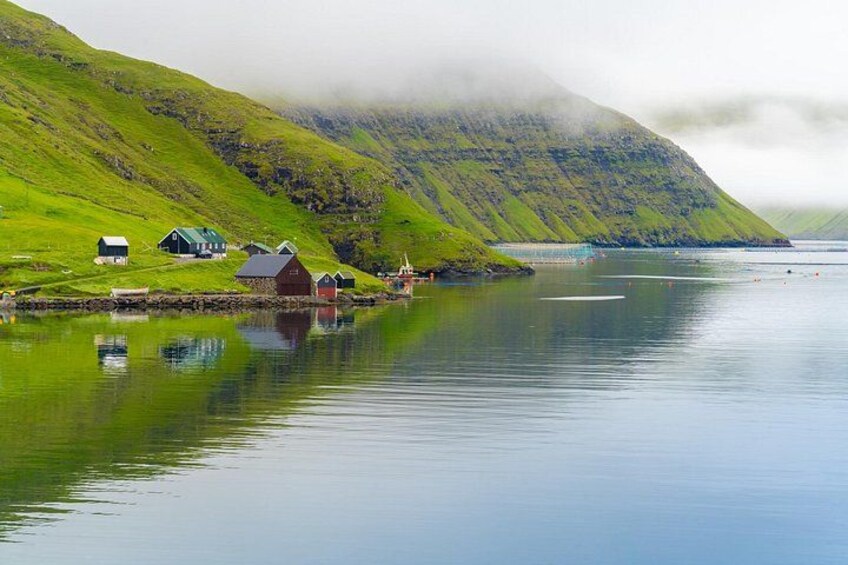 A calm lake with houses on the shore, surrounded by green hills and a misty mountain. 1