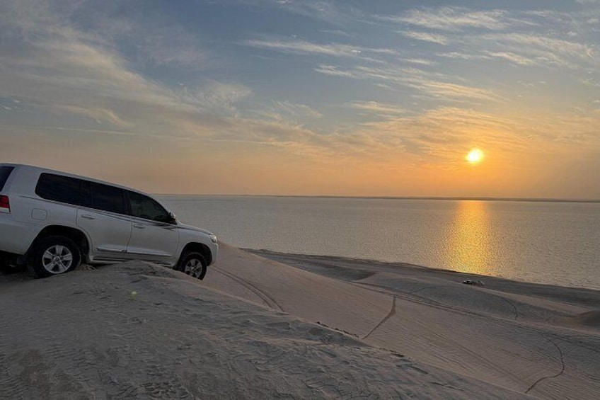 A white SUV parked on a sandy beach with the sun setting over the ocean. 2