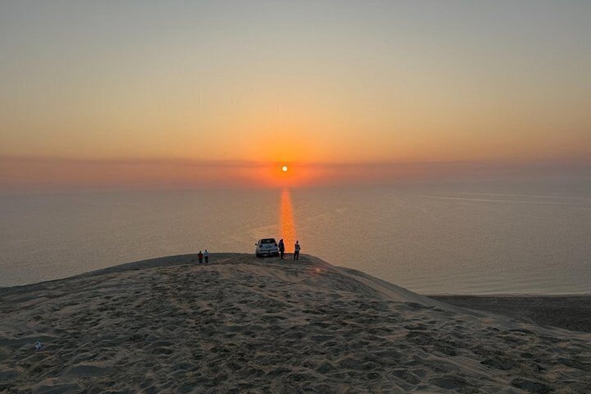 A sunset over a desert landscape with a dune, a vehicle, and a few people. 4