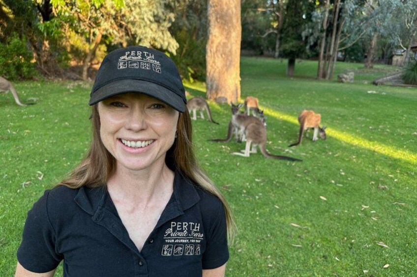 A person wearing a cap and a shirt with 'PERTH' written on it, standing in a grassy area with kangaroos in the background. 3