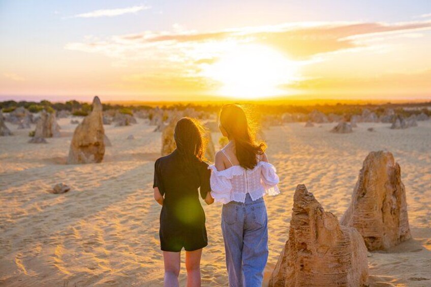 Two people standing on a sandy beach with rock formations in the background during sunset. 3