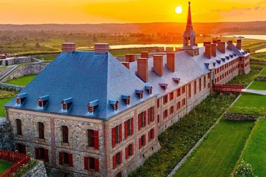 Un grand bâtiment en pierre, doté d’un toit en pente et de fenêtres rouges, entouré de champs verdoyants et bordé par une étendue d’eau. 1