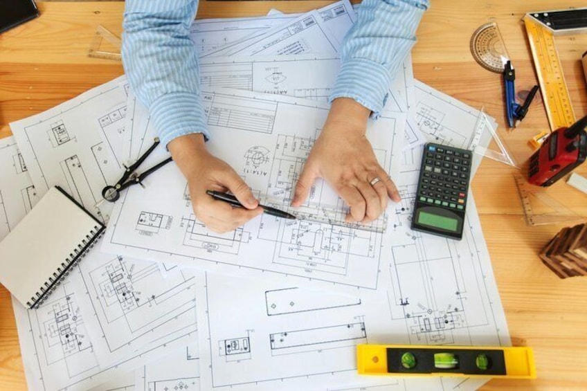 A person working with architectural plans, a calculator, and drafting tools on a wooden desk. 1