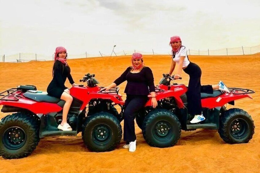 Three people on red ATVs in a desert with sand dunes and a fence in the background. 1