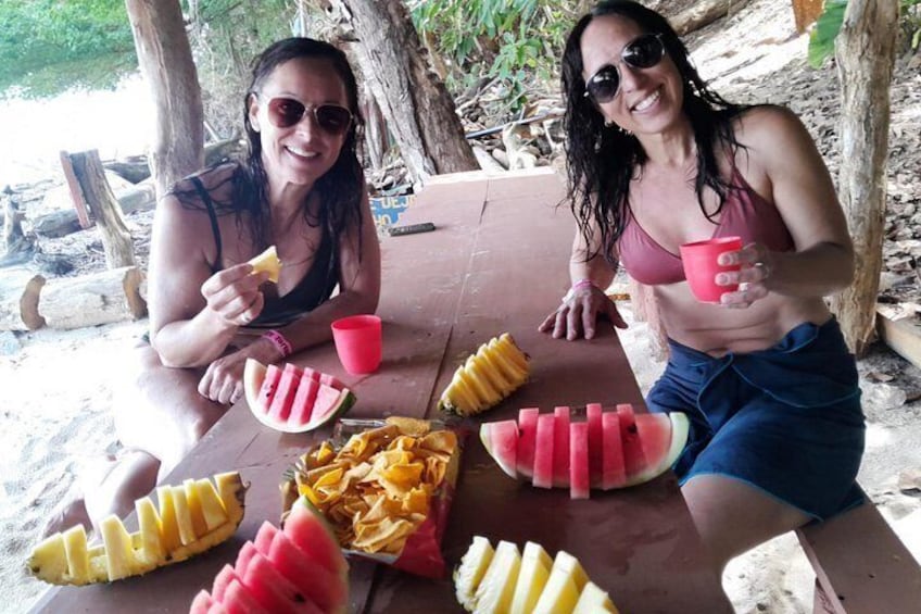 Two people sitting at a wooden table outdoors, eating watermelon and bananas, with a red cup on the table. 5