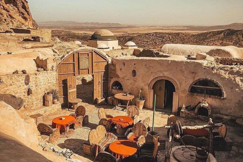 A traditional stone building with a wooden door, surrounded by outdoor seating and a view of a desert landscape. 4