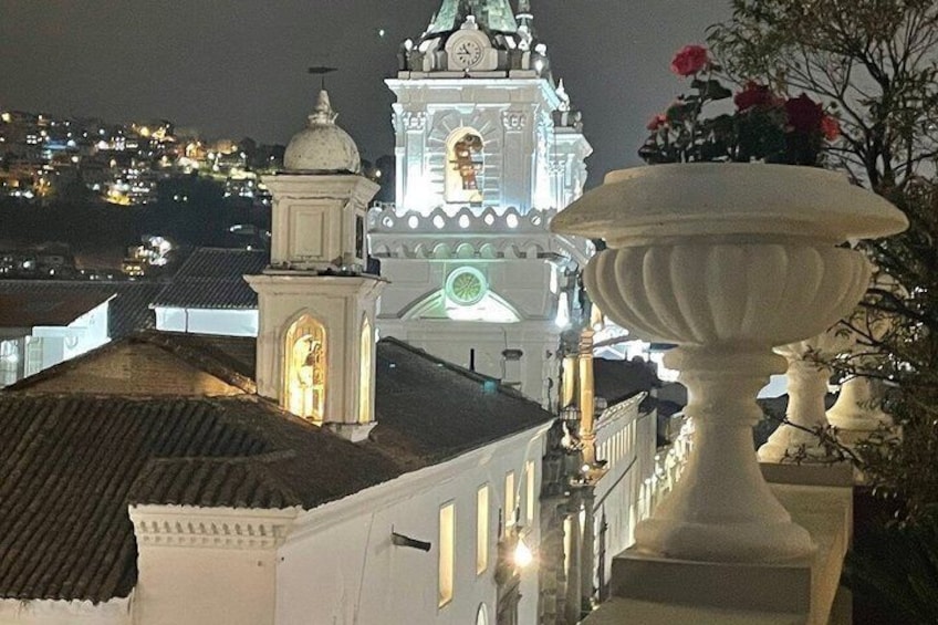 A night view of a church with illuminated clock and bell tower, a white urn, and a cityscape in the background. 2
